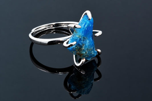 A silver-tone ring holds a rough bright blue crystal in curved arms on a dark reflective surface.