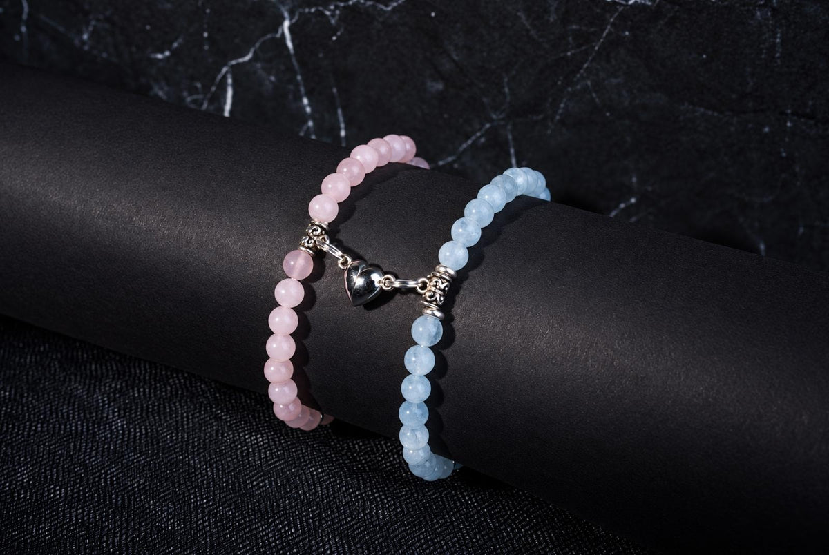 Two beaded bracelets, one pink and one pale blue, with a silver heart charm, on a dark cylindrical surface.