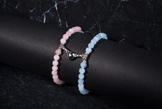 Two beaded bracelets, one pink and one pale blue, with a silver heart charm, on a dark cylindrical surface.