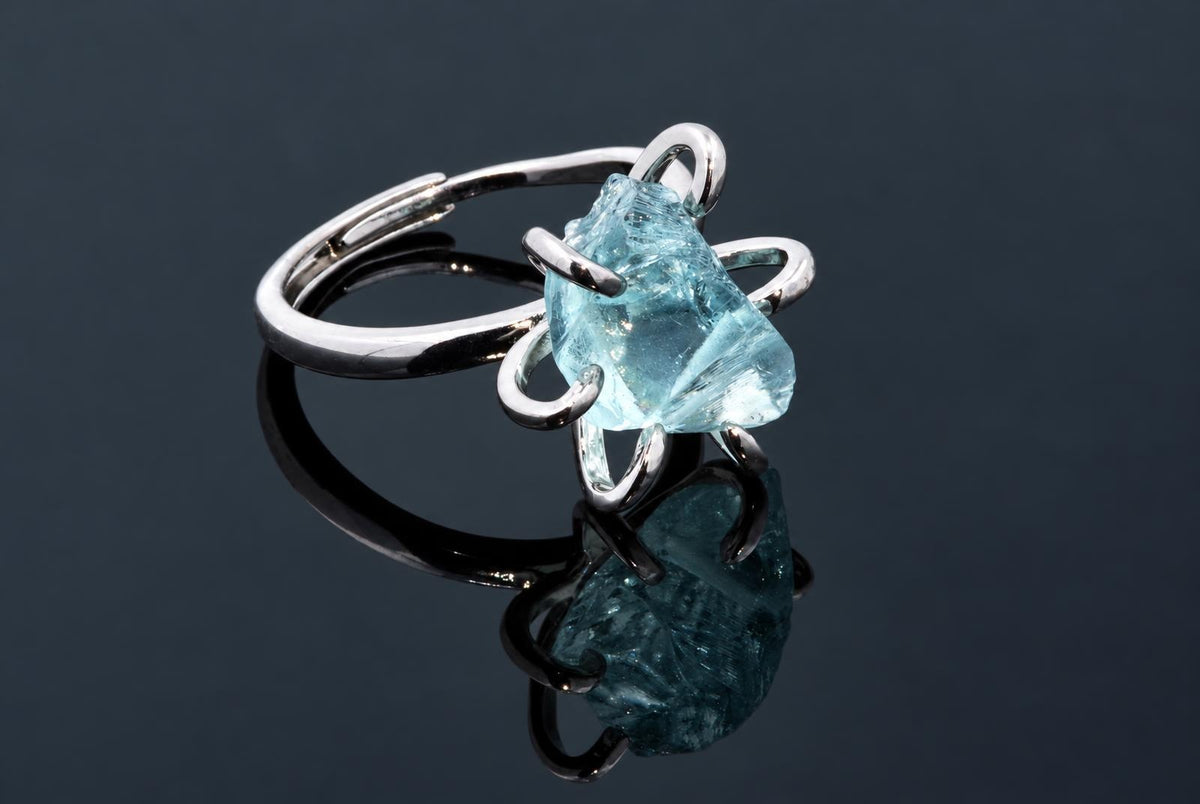 A silver-tone ring holds a rough pale blue crystal in curved arms on a dark reflective surface.