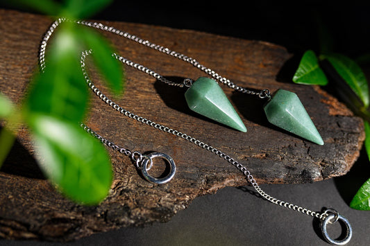 Green faceted pendulums on silver-tone chains lie on a wooden surface among green leaves.