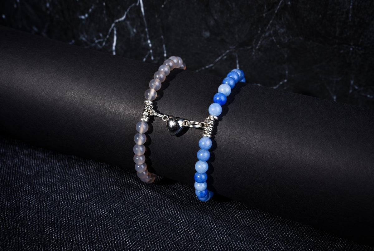 Two beaded bracelets on a dark surface: one with pale beads and a silver heart charm, the other with blue beads.