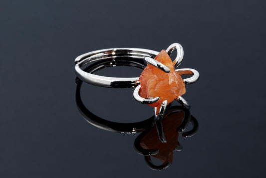 A silver-tone ring holds a rough orange crystal in curved arms on a dark reflective surface.