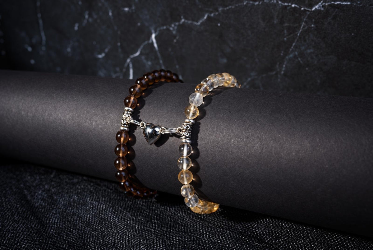 Two beaded bracelets on a dark cylindrical form: one with brown beads, the other with clear and yellow beads. A silver heart charm connects them.