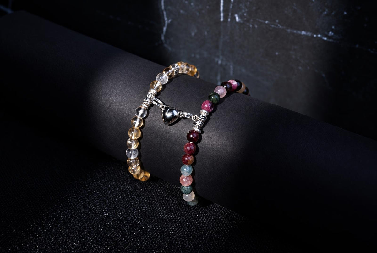 Two beaded bracelets on a dark surface; one has yellow and clear beads with a silver heart charm, the other has multicoloured red, green and pink beads.