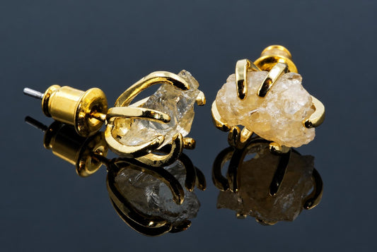 A pair of gold-tone stud earrings with raw, uncut stones in a claw setting on a dark reflective surface.
