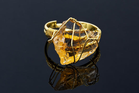 A gold-coloured ring holds a rough transparent yellow crystal wrapped in thin wire on a dark reflective surface.