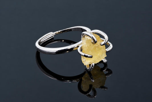 A silver-tone ring holds a rough yellow crystal in curved arms on a dark reflective surface.