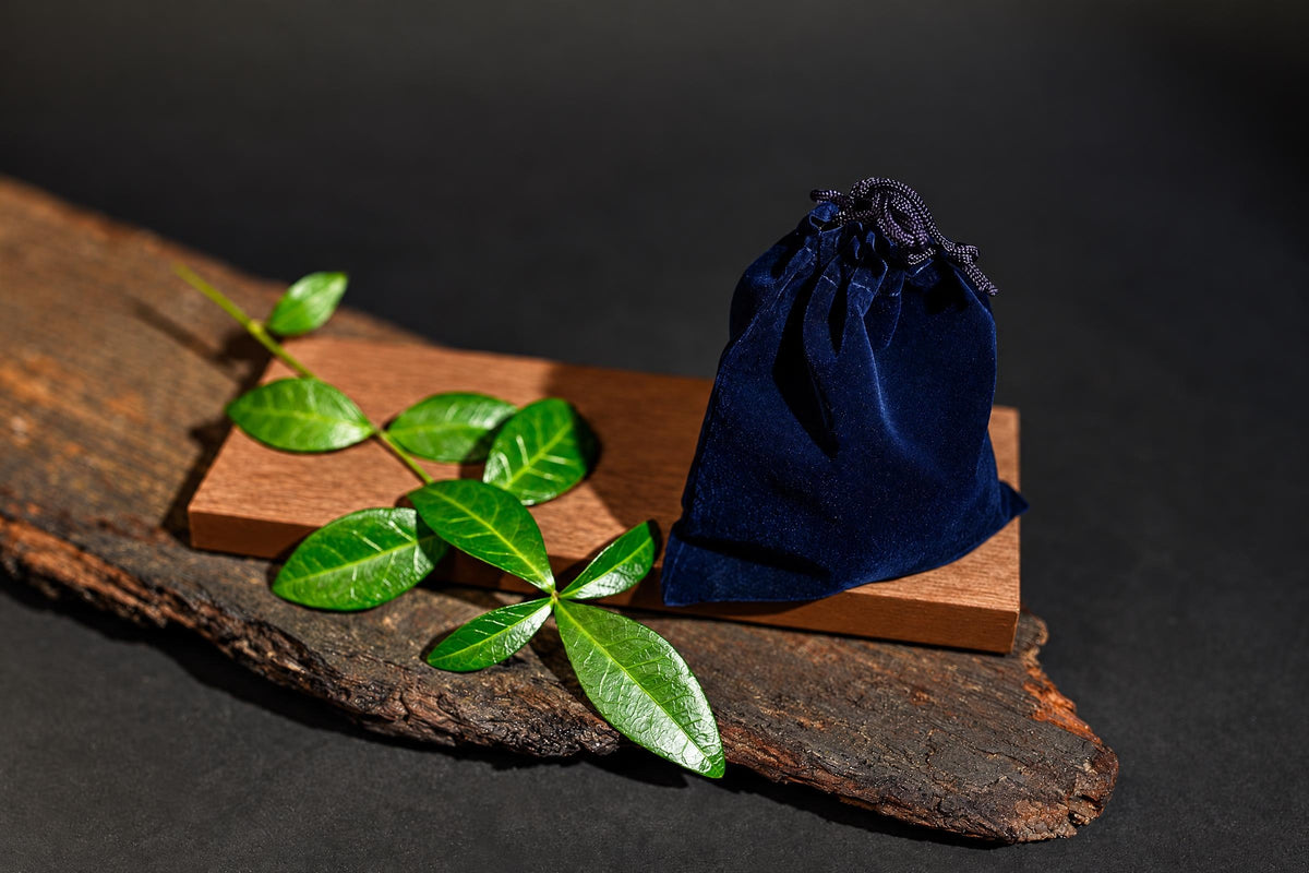 A dark blue velvet pouch with a drawstring sits on a wooden block, next to green leaves on a rustic wood slice.
