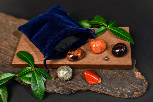 A blue velvet pouch sits on a wooden board with several polished stones and green leaves nearby.