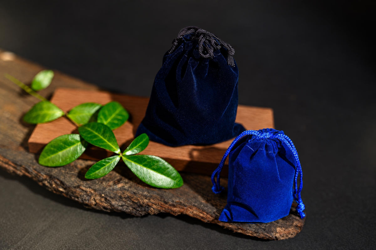 Two blue velvet drawstring pouches sit on a dark wooden surface with green leaves and a small wood block.