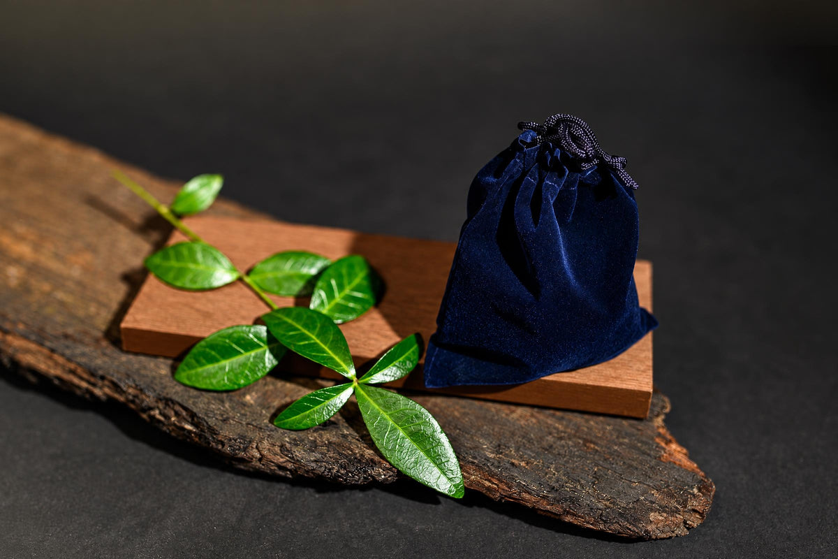 A dark blue velvet pouch with a drawstring sits on a wooden block beside green leaves on a rustic wood slab.