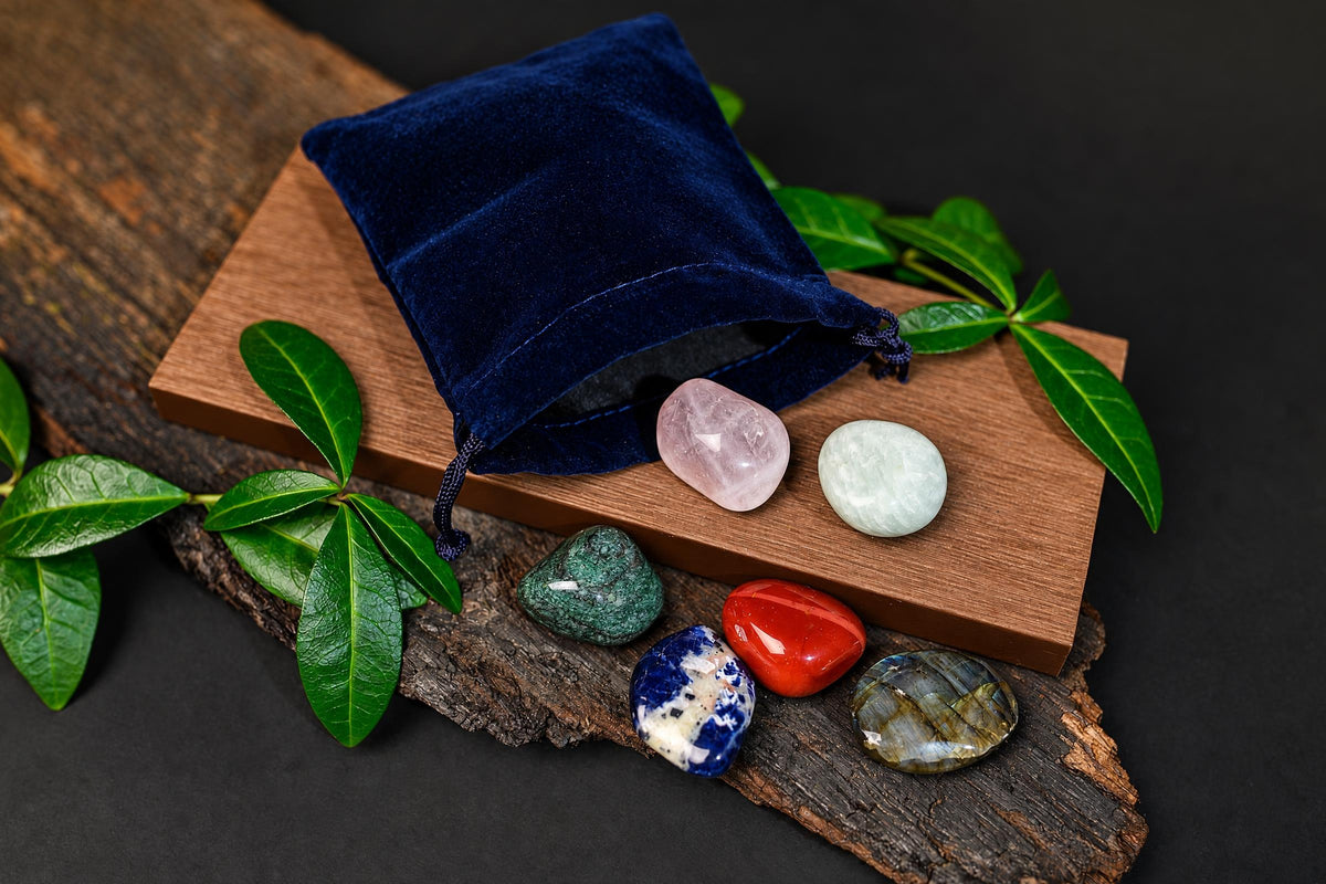 An open navy-blue pouch sits beside colourful polished stones on a wooden surface among green leaves.
