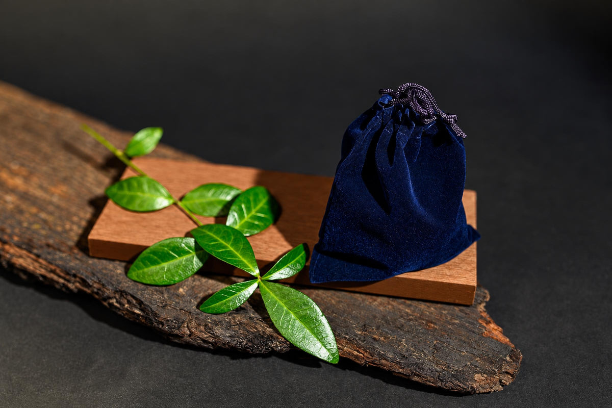 A navy-blue drawstring pouch stands on a wooden surface beside green leaves against a dark background.