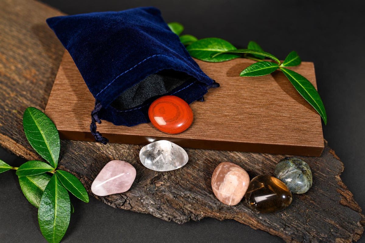 A dark blue velvet pouch sits on a wooden board with a red stone. Five polished stones in pink, clear, peach, brown and green rest on a dark wood surface beside green leaves.