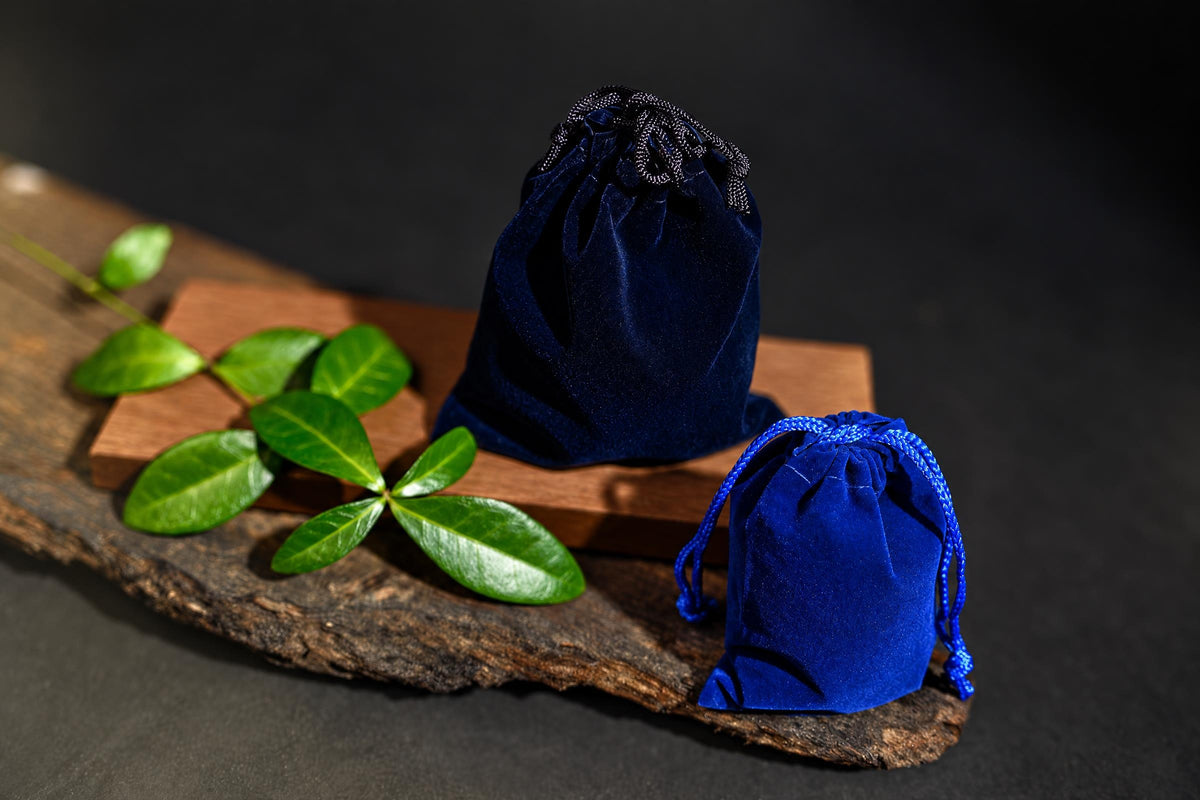 Two blue velvet drawstring pouches sit on a wooden surface with green leaves nearby.