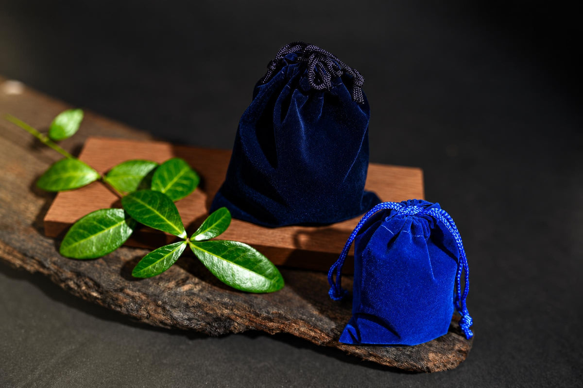 Two blue velvet drawstring pouches sit on a wooden surface with green leaves nearby.