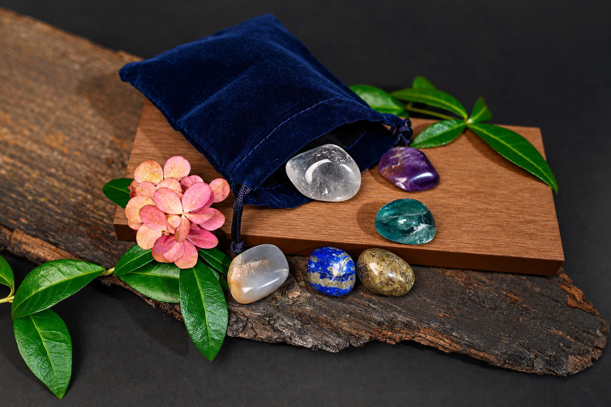 A dark blue velvet pouch rests on a wooden board with seven polished stones in clear, purple, green, white, and blue. A pink flower and green leaves decorate the scene.