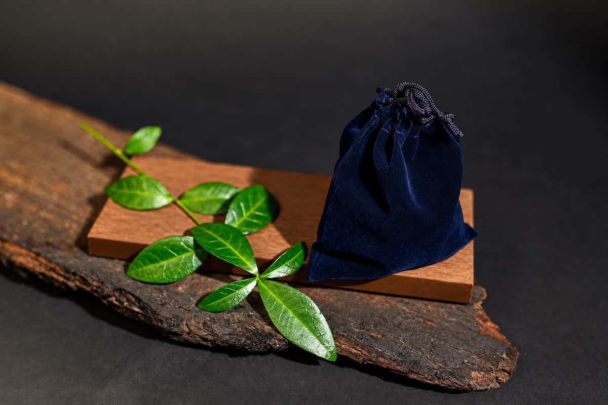 A dark blue velvet pouch with a drawstring sits on a wooden block, next to green leaves on a rustic wood slab.