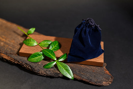 A dark blue velvet pouch with a drawstring sits on a wooden block, next to green leaves on a rustic wood slab.