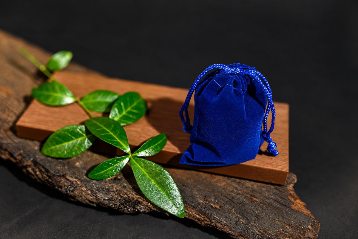 A blue velvet pouch with a drawstring sits on a wooden surface next to green leaves.