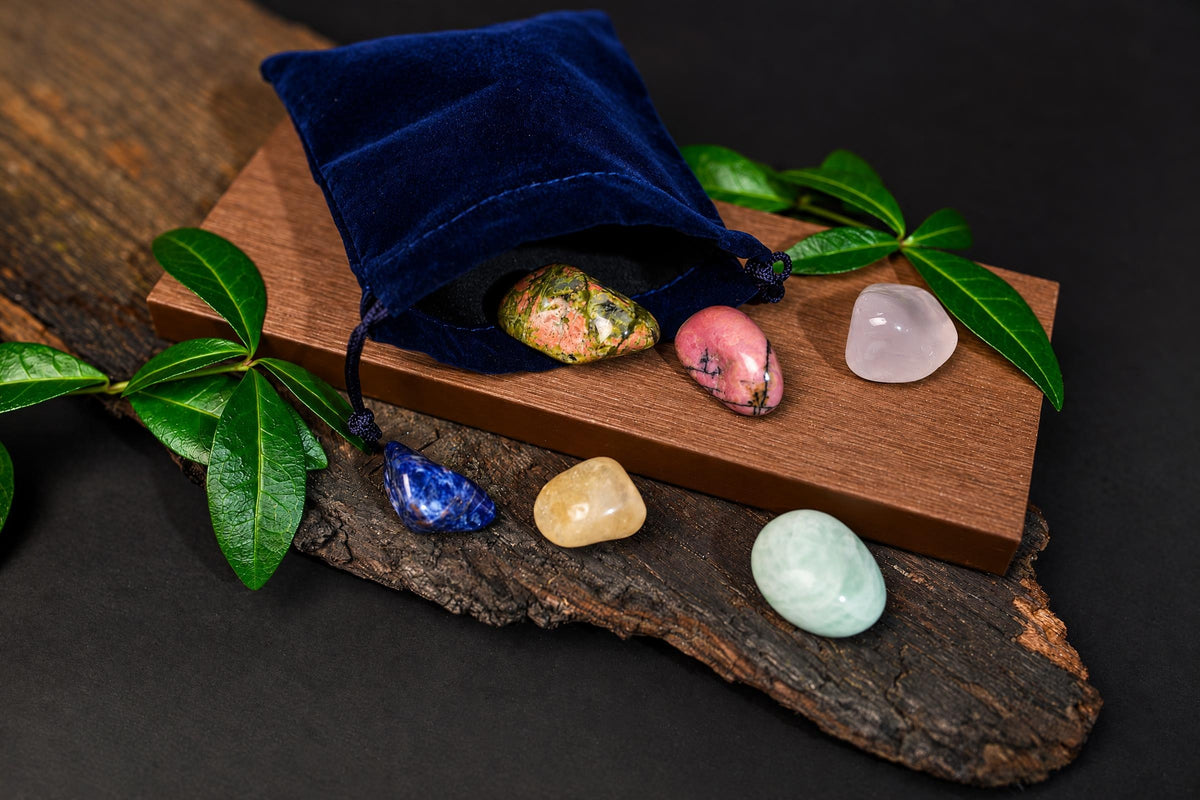 An open navy-blue pouch and polished pink, blue, pale green and patterned stones rest on a wooden surface among green leaves.
