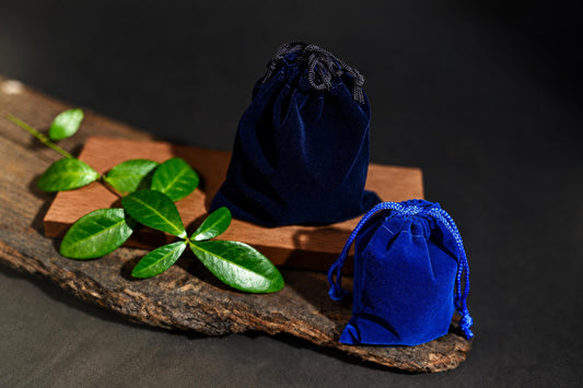 Navy-blue and bright blue drawstring pouches rest on a wooden surface beside green leaves against a dark background.