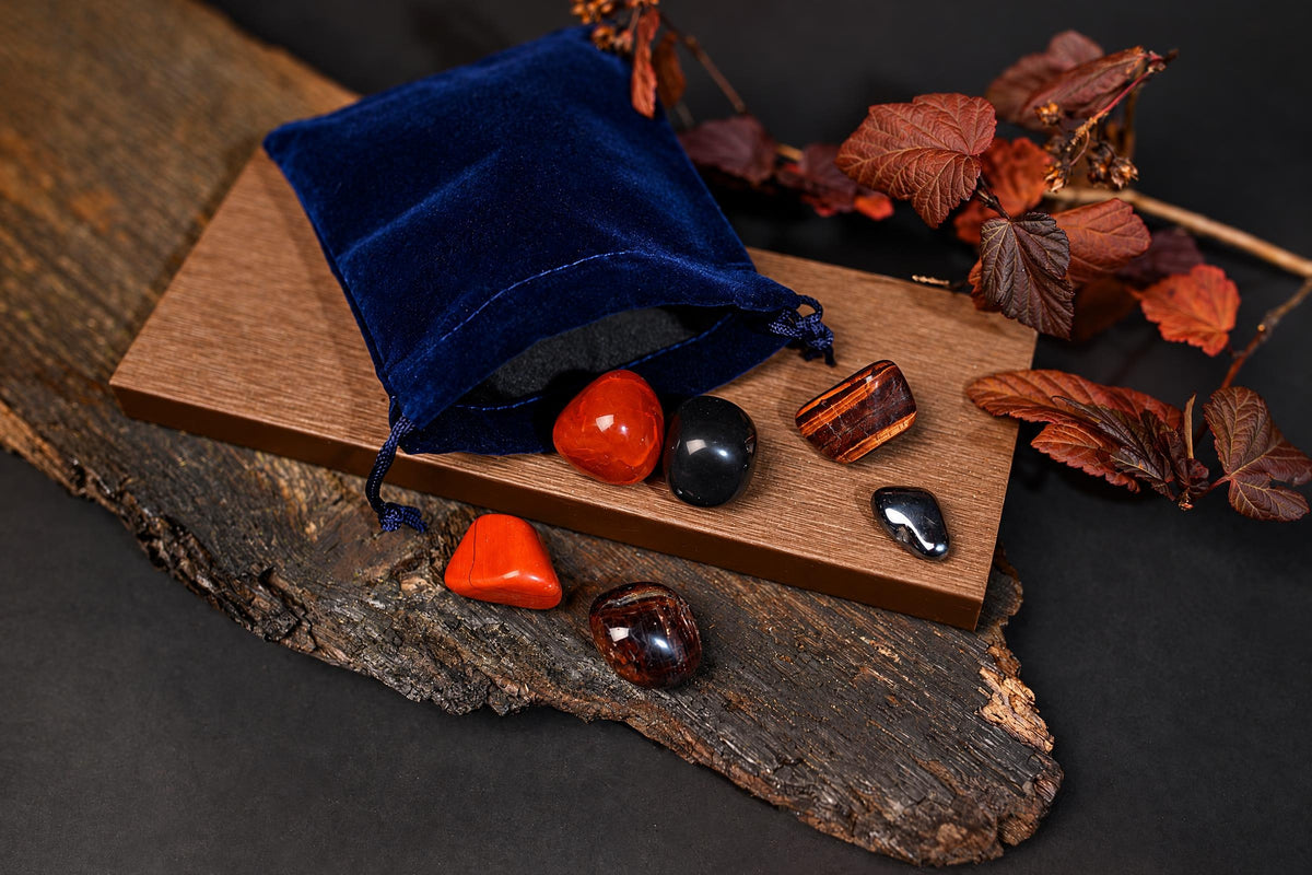 A blue velvet pouch sits on a wooden board with several polished stones in red, black, and striped patterns, surrounded by dried leaves.