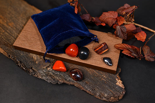 A blue velvet pouch sits on a wooden board with several polished stones in red, black, and striped patterns, surrounded by dried leaves.