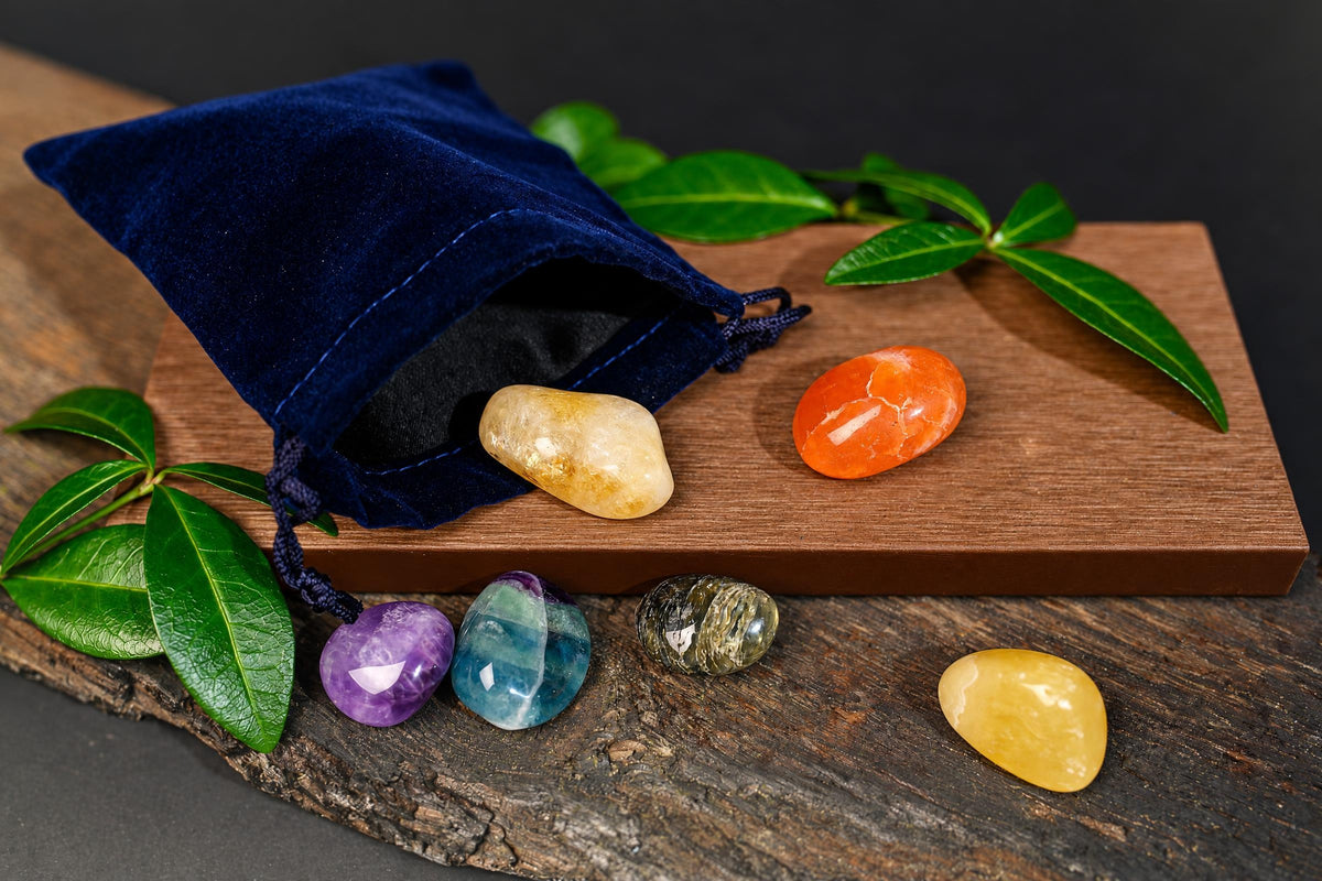 A blue velvet pouch with drawstrings sits on a wooden surface, surrounded by six polished stones in purple, blue, green, orange, yellow, and cream tones.