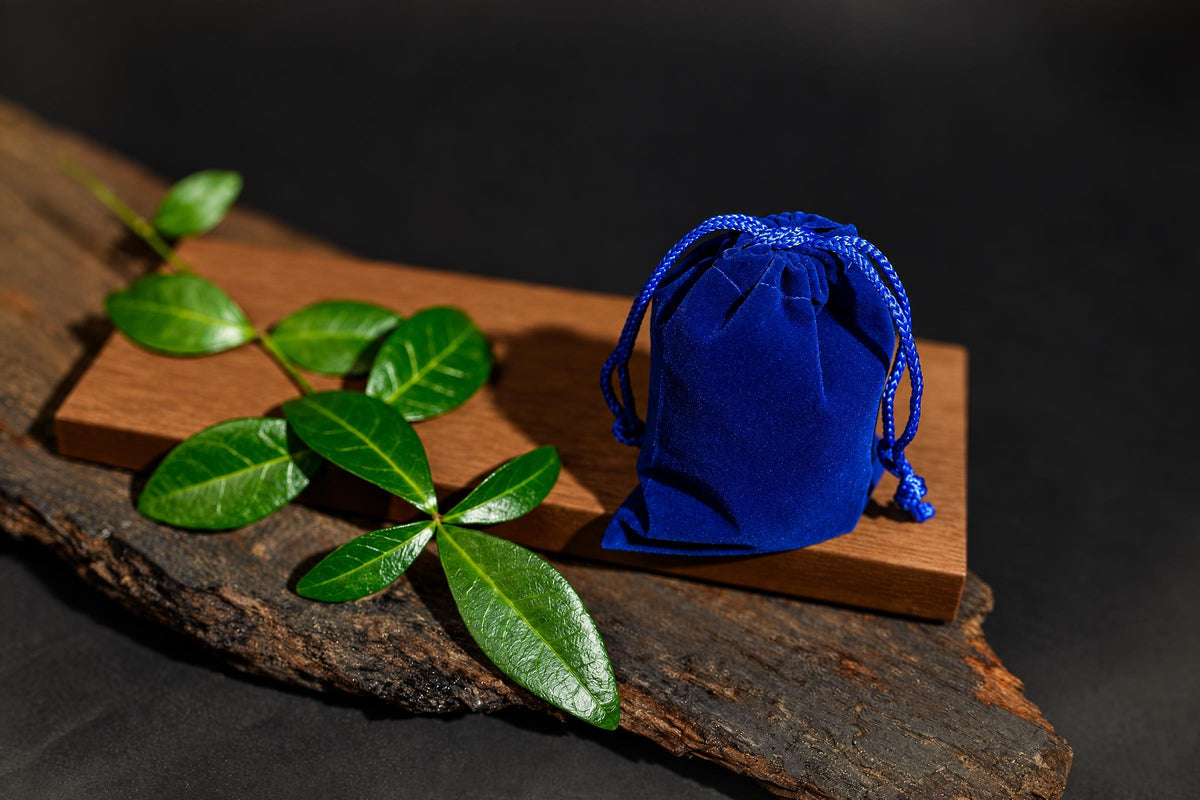 A blue velvet drawstring pouch sits on a wooden board with green leaves, against a dark background.