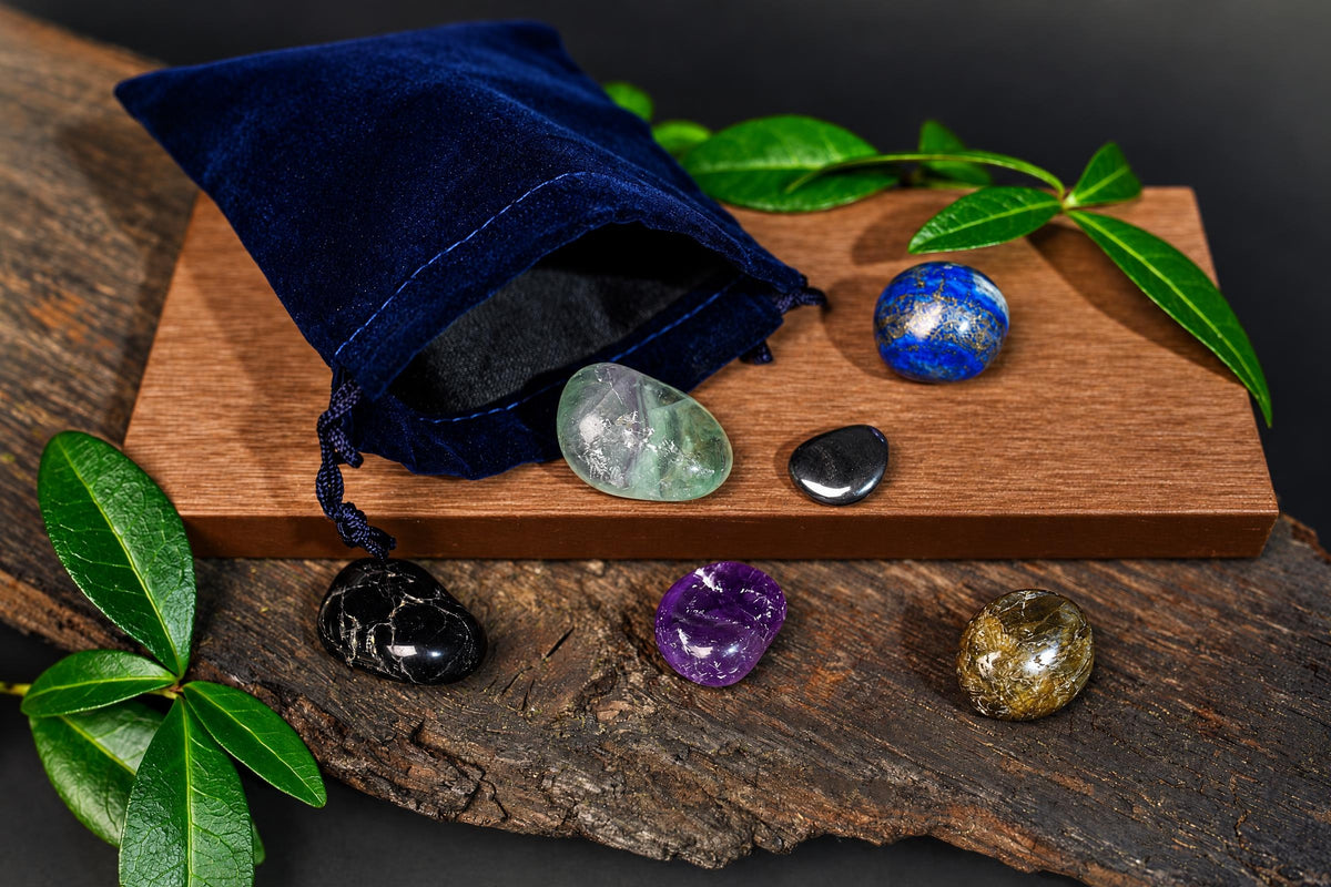 An open navy-blue pouch and polished blue, green, purple and black stones rest on a wooden surface among green leaves.