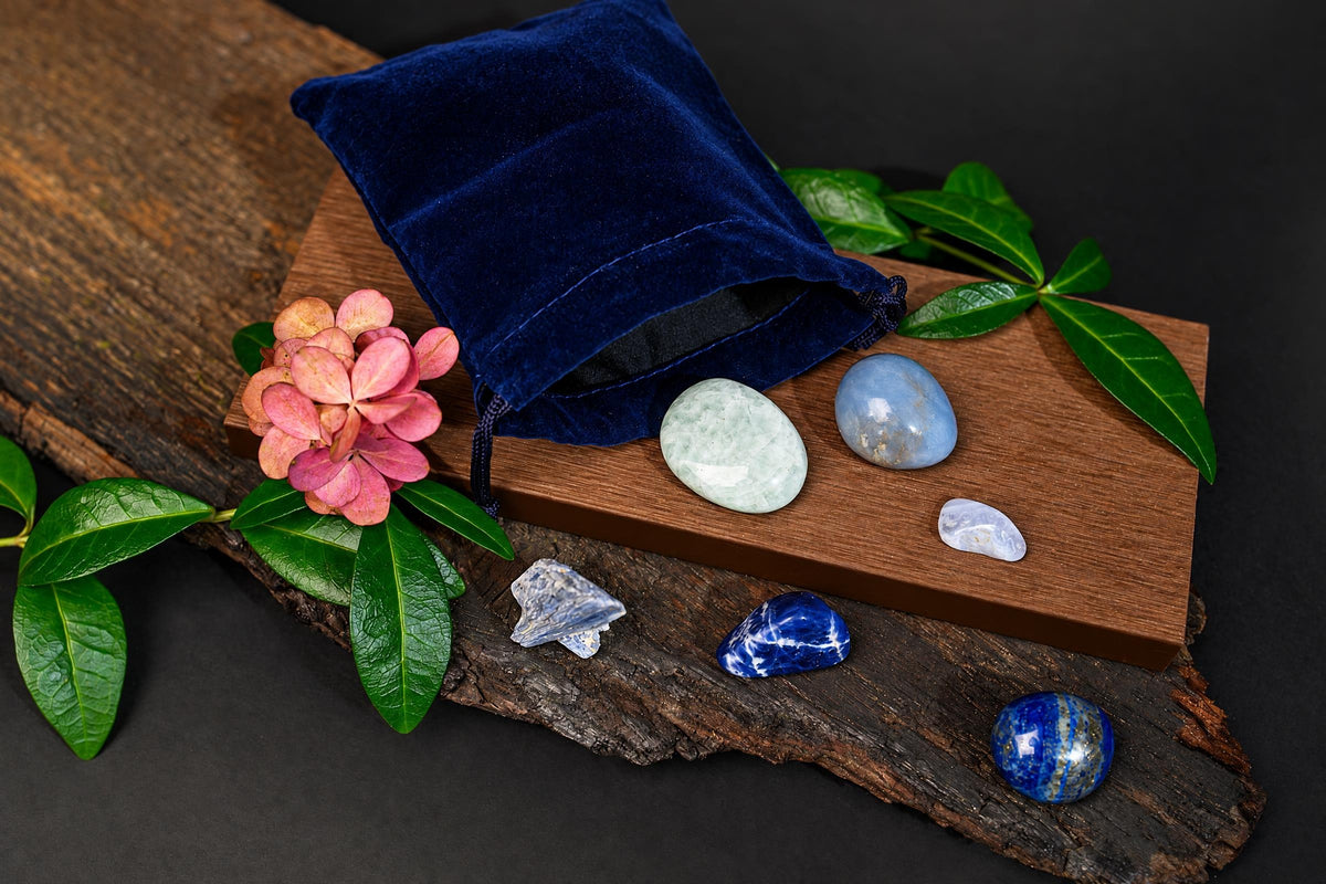 A blue velvet pouch sits on a wooden board with several polished stones and crystals, including green, blue, and white pieces, arranged on wood and dark surfaces.