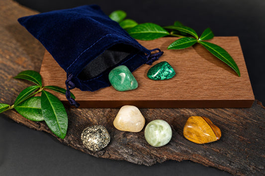 A blue velvet pouch sits on a wooden board with green leaves, surrounded by six polished stones in various colours on wood.