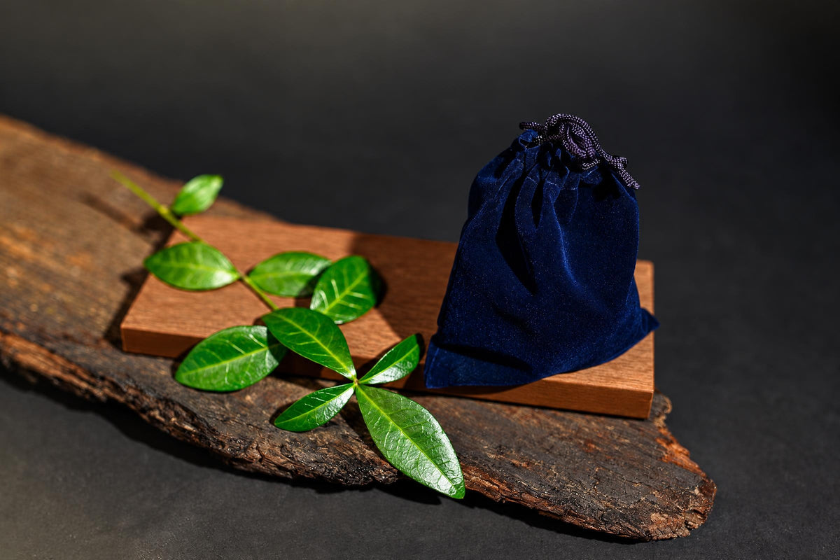 A dark blue velvet drawstring pouch sits on a wooden block, next to green leaves on a rustic wood slab.
