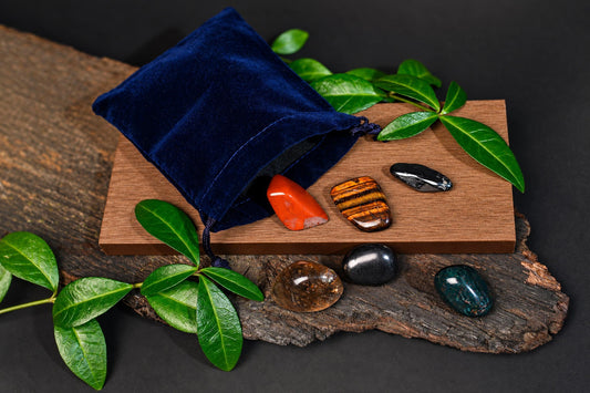 A blue velvet pouch spills polished stones onto a wooden board and dark surface, surrounded by green leaves.