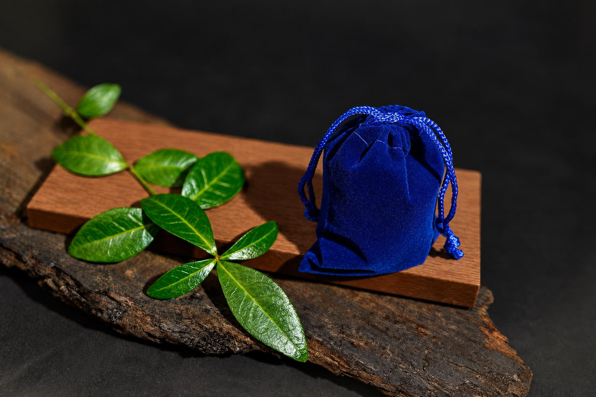 A blue velvet pouch with a drawstring sits on a wooden board next to green leaves.