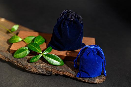 Two blue velvet drawstring pouches sit on a wooden surface with green leaves and a dark background.