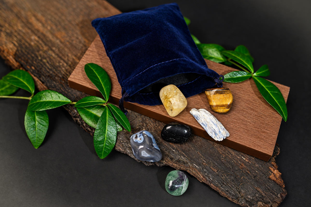 A dark blue velvet pouch sits on a wooden board with several polished stones and green leaves nearby.