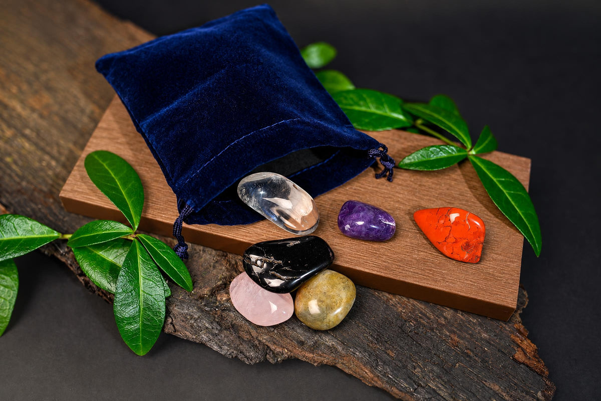 A blue velvet pouch sits on a wooden board with six polished stones in clear, purple, orange, black, pink and yellow-green hues.