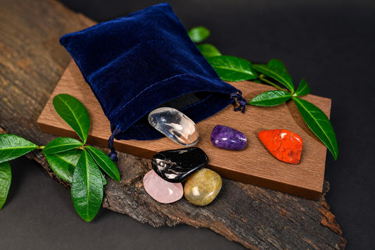 A blue velvet pouch sits on a wooden board with six polished stones in clear, purple, orange, black, pink and yellow-green hues.