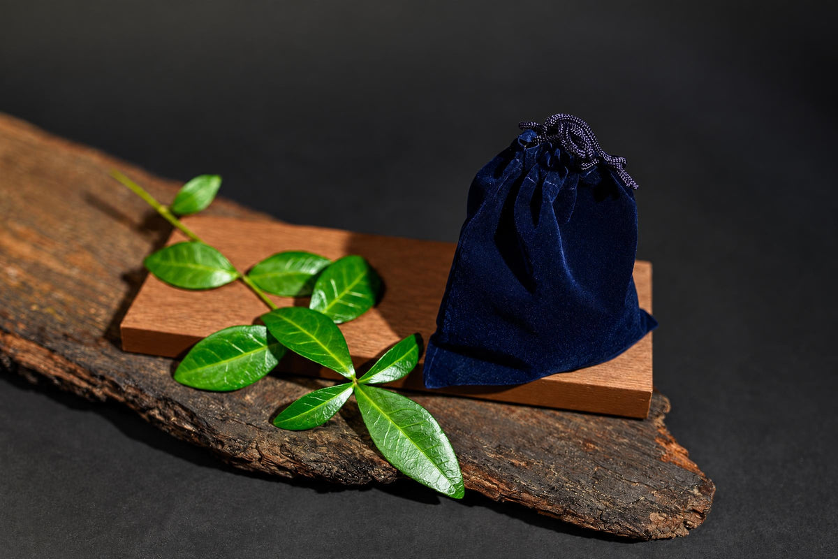 A dark blue velvet pouch with a drawstring sits on a wooden block, next to green leaves on a rustic wood slab.