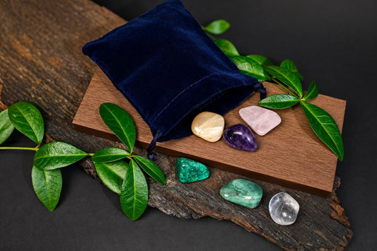 A dark blue velvet pouch rests on a wooden board, spilling out seven polished stones in cream, pink, purple, green, and clear hues.