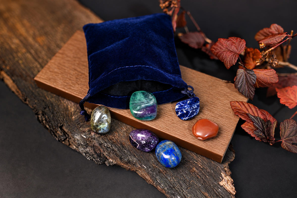 A blue velvet pouch sits on a wooden board with six polished stones in green, purple, blue, and orange.