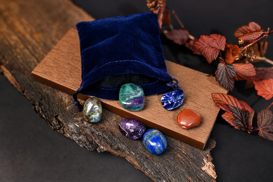 A blue velvet pouch sits on a wooden board with six polished stones in green, purple, blue, and orange.