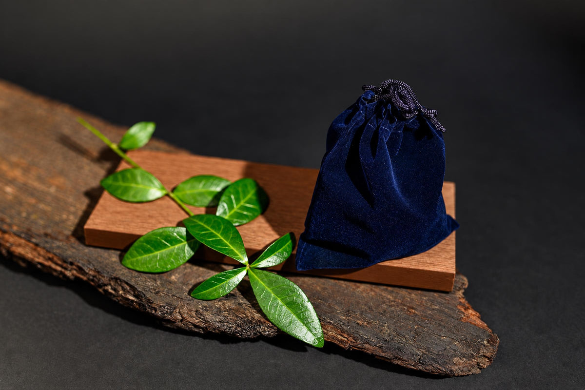 A dark blue velvet pouch with a drawstring sits on a wooden block next to green leaves on a rustic wood slab.