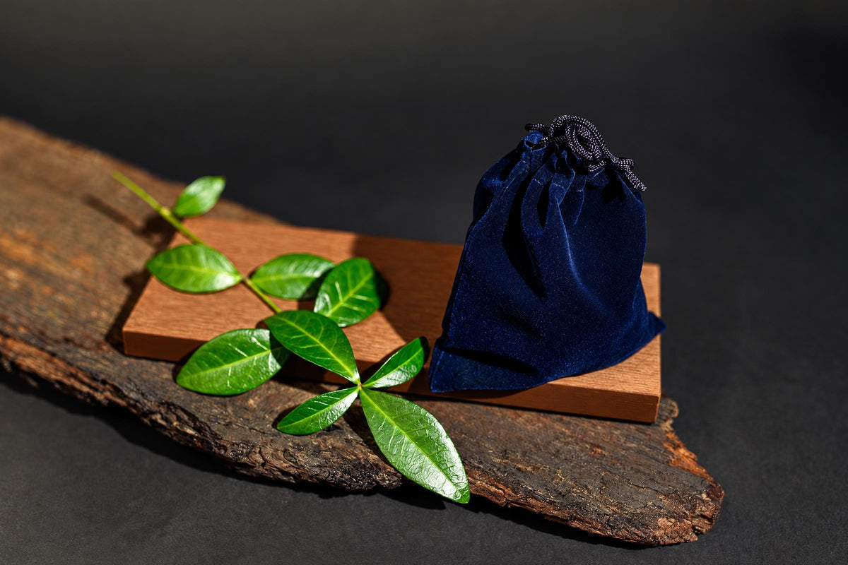 A closed dark blue drawstring pouch stands on a wooden board among green leaves against a dark background.