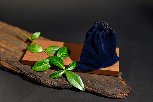 A closed dark blue drawstring pouch stands on a wooden board among green leaves against a dark background.