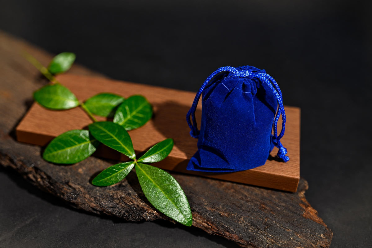 A blue velvet drawstring pouch sits on a wooden board with green leaves, all on a dark surface.