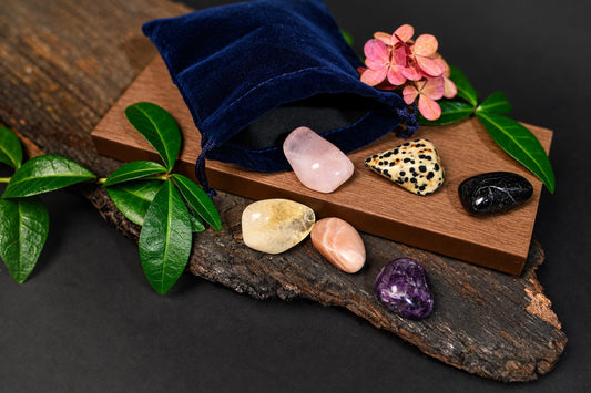 A blue velvet pouch sits on a wooden board with seven polished stones, pink flowers, and green leaves on a dark surface.